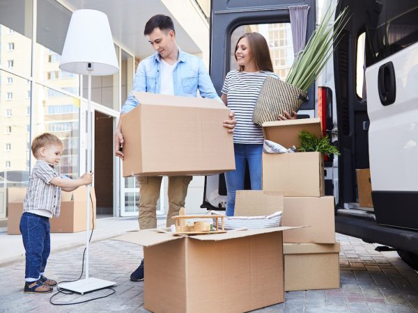 Portrait of happy young family holding cardboard boxes standing next to moving van and smiling looking at their son